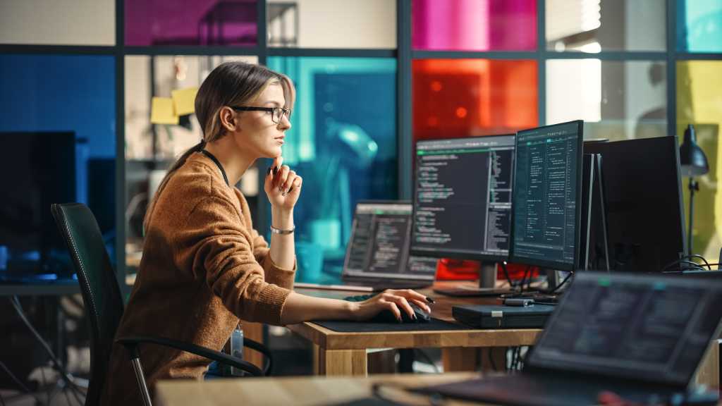 Female Software Engineer Writes Code on Desktop Computer With Two Monitors and Laptop Aside In Stylish Office. Caucasian Woman Working On Artificial Intelligence Service For Big Tech Company.